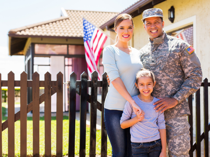 A man in his military uniform, a woman, and their child stand in front of their house.