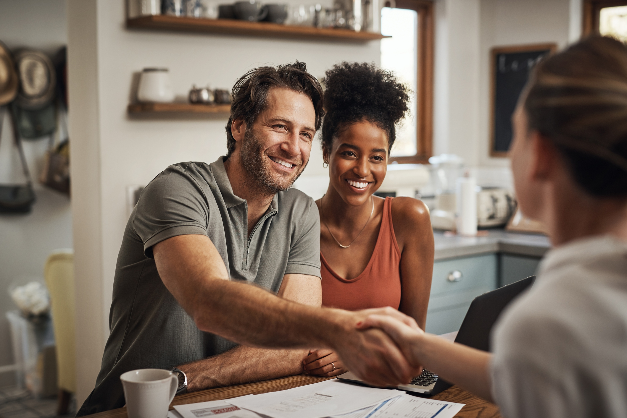 A young man and woman shake hands with their real estate agent at their kitchen table.